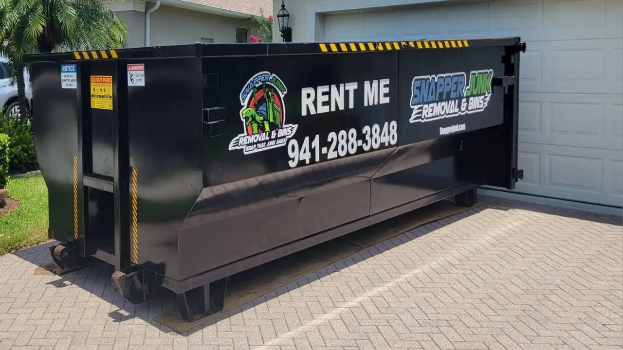 A black Snapper roll-off lettered RENT ME 941-288-3848 with the turtle roundel and SNAPPER JUNK wordmark, parked on herringbone pavers in front of a garage door