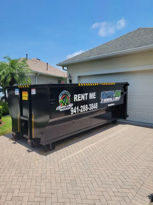 Snapper's black roll-off lettered RENT ME 941-288-3848 on a herringbone paver driveway beside a palm